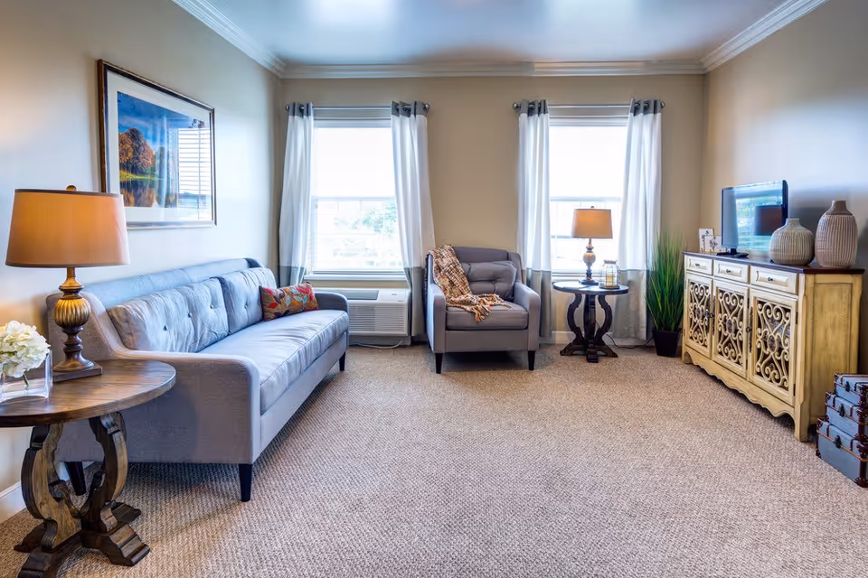 A bright and cozy living room with beige walls and carpet. The room features a gray sofa with a colorful pillow, a matching gray armchair with a throw blanket, two wooden side tables each with a lamp, a decorative cabinet with a flat-screen TV on top, and two large windows with white curtains letting in natural light.