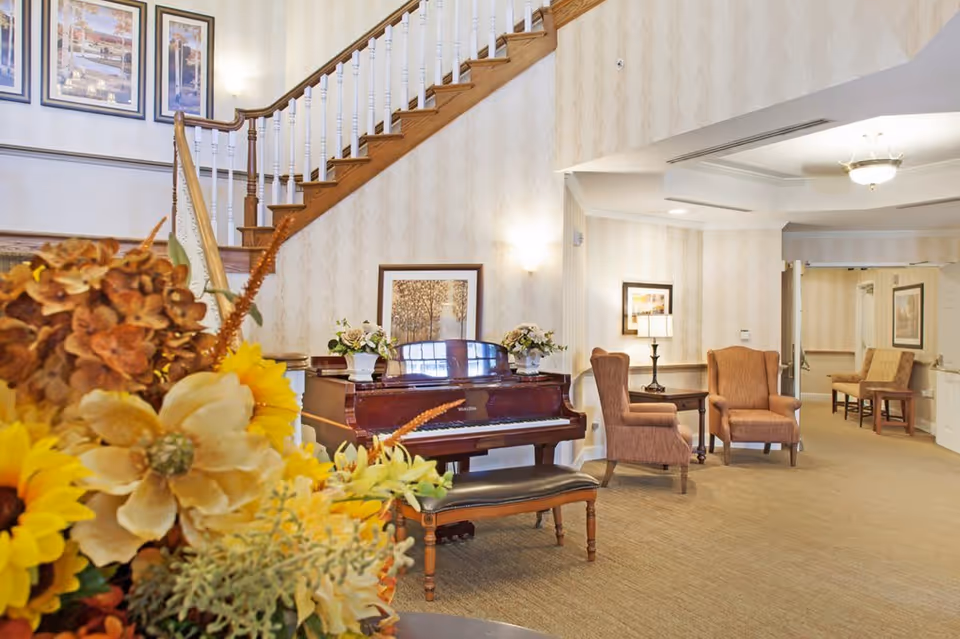 A bright and welcoming interior common area of Poland Village Assisted Living featuring a grand piano with a bench, two upholstered armchairs with a small table and lamp between them, floral arrangements, framed artwork on the walls, and a staircase with wooden handrails and white balusters.
