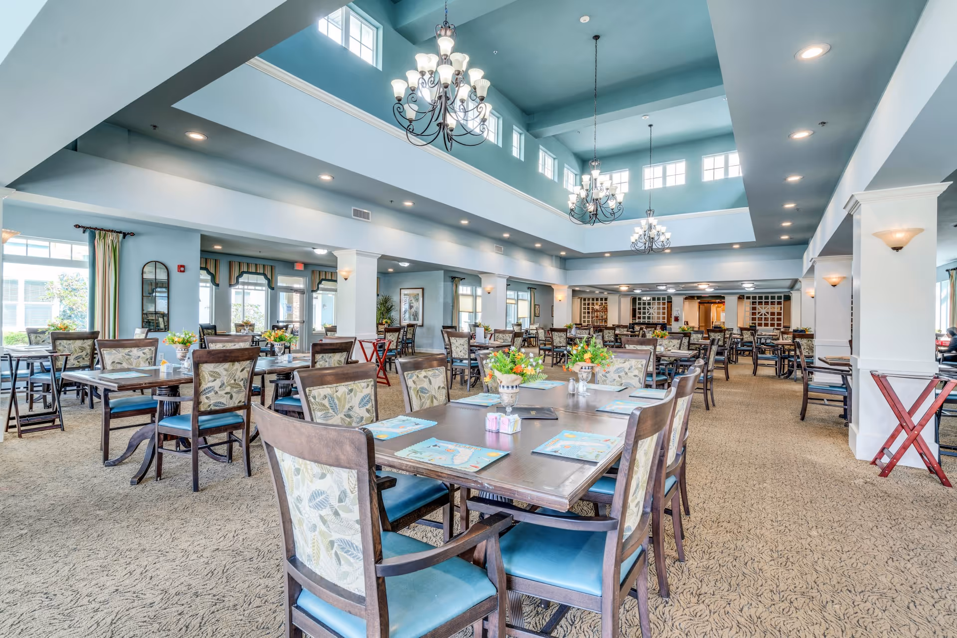 Spacious, brightly lit dining room with multiple set tables, chandeliers, and a two-story ceiling in a senior living facility.
