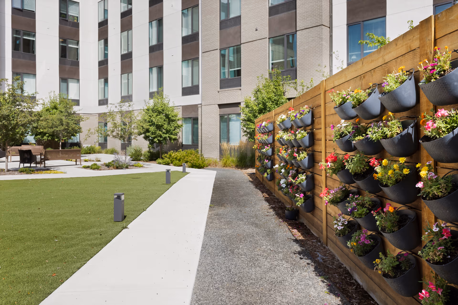 Outdoor garden area at Hyland Hills Senior Living featuring a pathway alongside a wooden fence with multiple black flower pots filled with colorful flowers. The background shows a multi-story building with windows and some trees and benches in a landscaped courtyard.