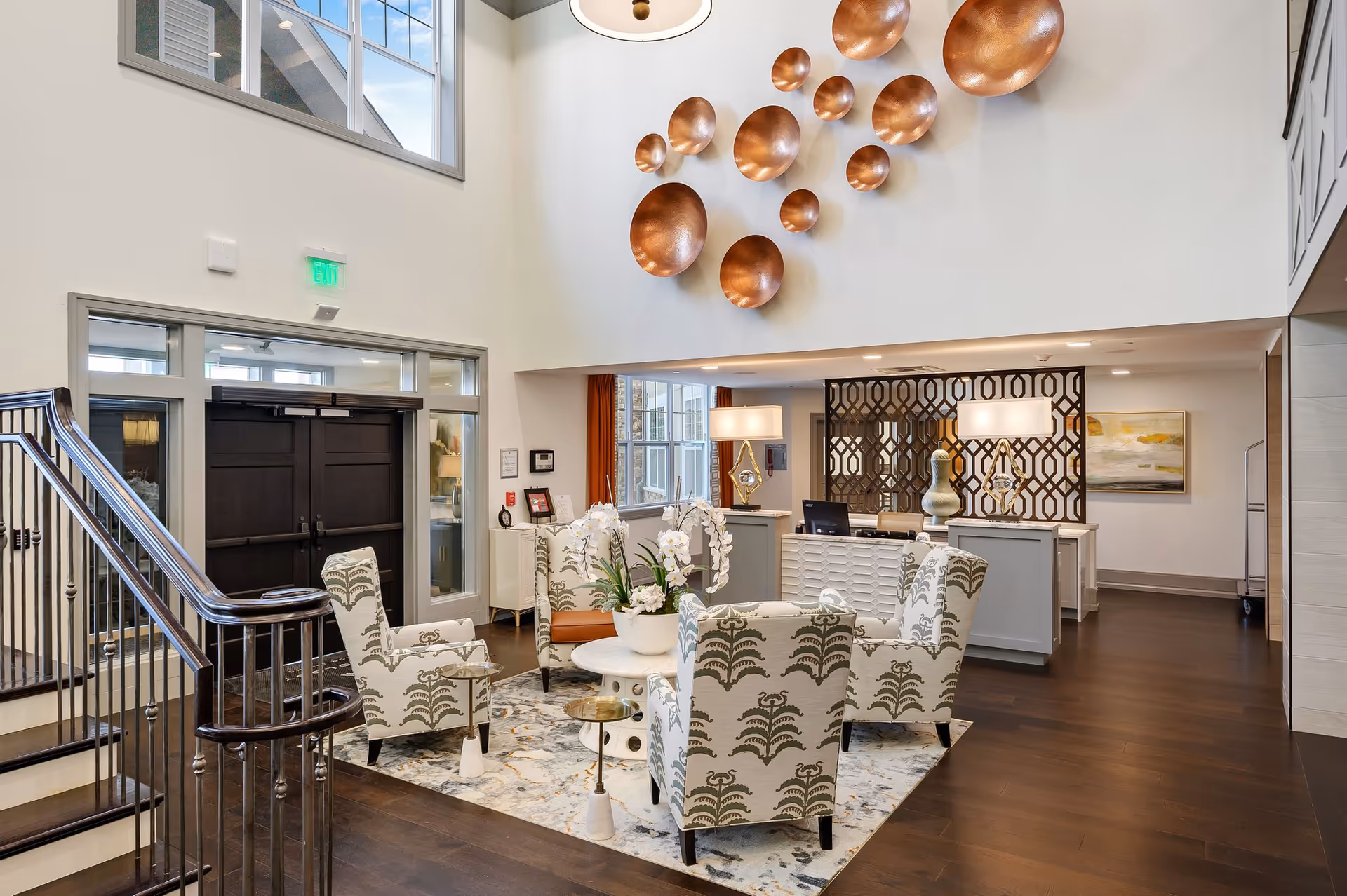 A bright and spacious lobby area with high ceilings featuring a decorative wall installation of various sized copper bowls. The seating area includes four patterned armchairs arranged around a round white table with a white orchid centerpiece on a patterned rug. Behind the seating area is a reception desk with two lamps and decorative items, and a geometric room divider. To the left is a staircase with a metal railing and double dark wooden doors with glass panels above and beside them.