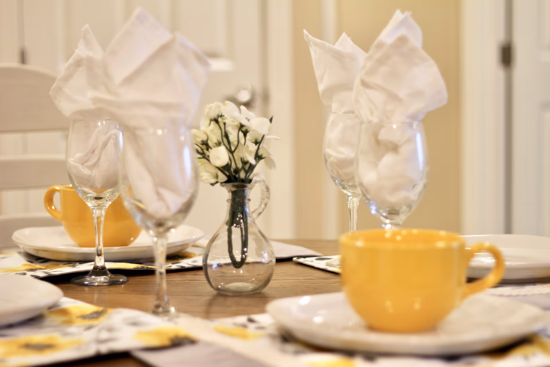Close-up view of a dining table set with white plates, yellow cups, wine glasses with white folded napkins, and a small glass vase with white flowers in the center.