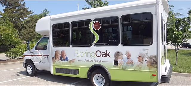 A white and green shuttle bus parked outdoors with the logo and name 'Spring Oak Assisted Living' on the side, featuring images of elderly people smiling and interacting.