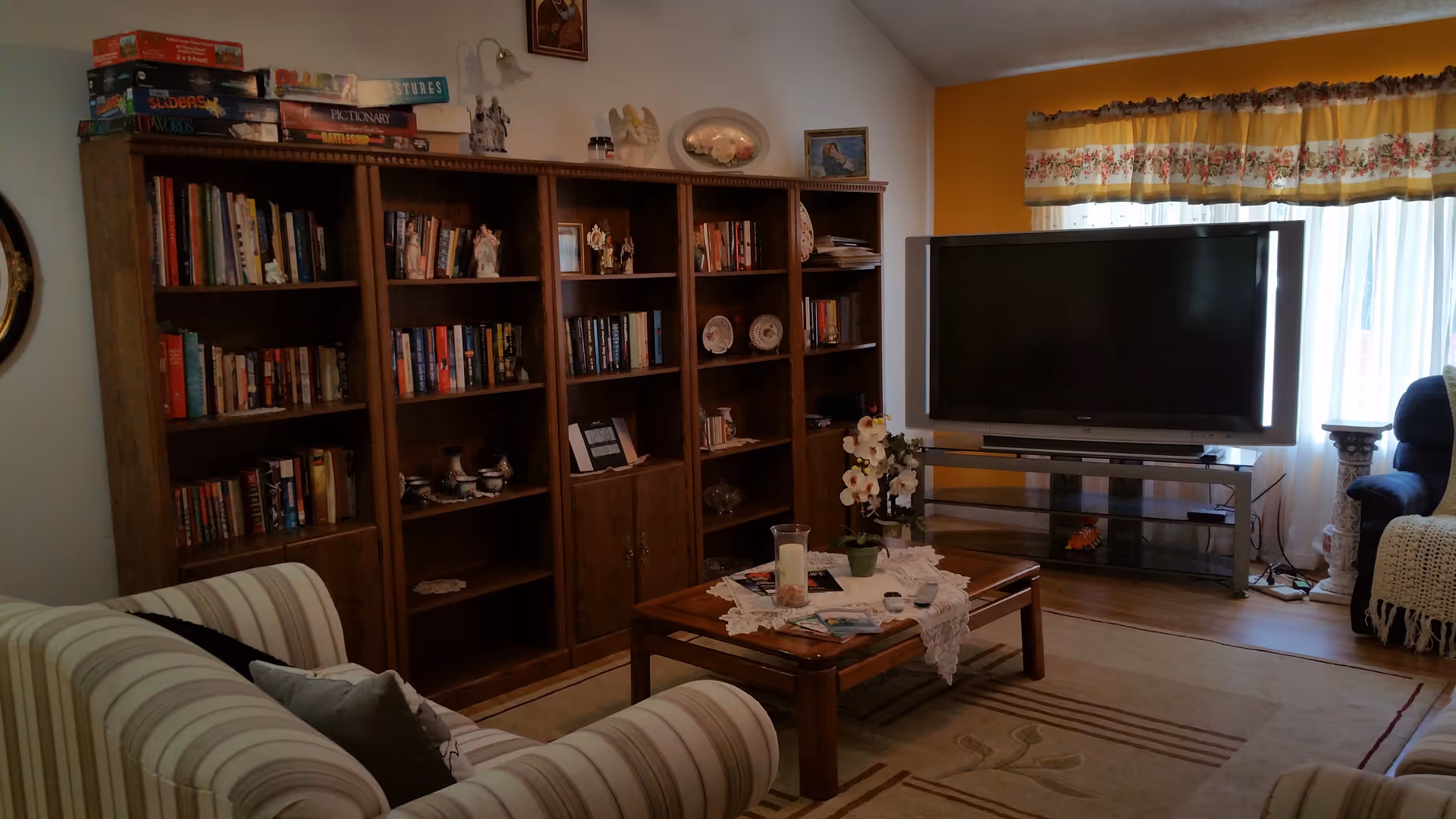 A cozy living room with a striped sofa, a wooden coffee table with a lace runner and decorative items, a large wooden bookshelf filled with books and ornaments, and a flat-screen TV on a glass stand in front of a window with floral curtains.