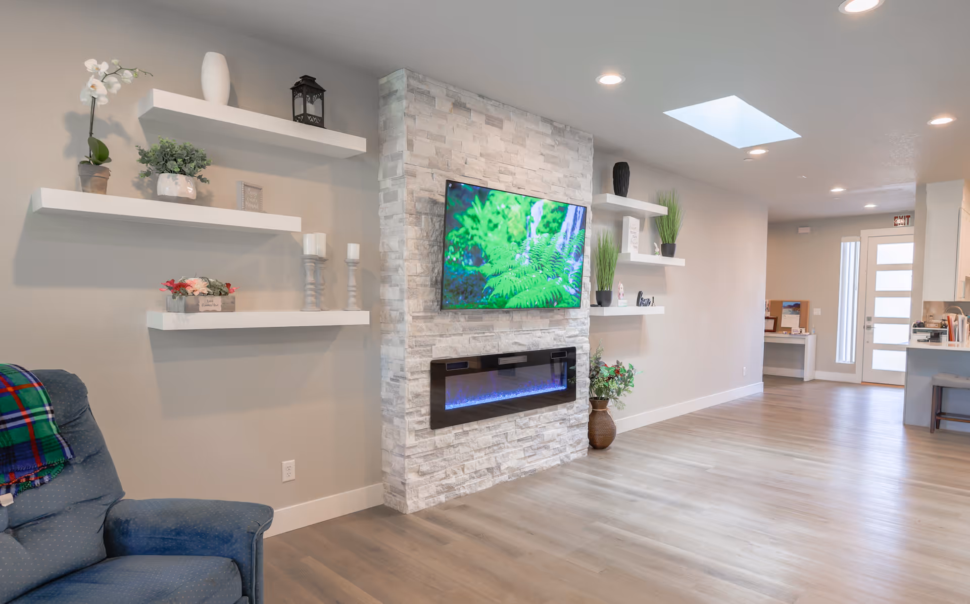 Bright modern living room with a wall-mounted TV above a stone fireplace, floating shelves, and a recliner.