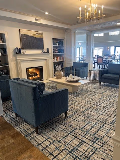 A cozy senior living facility common area with a lit fireplace, blue upholstered armchairs arranged around a white coffee table, built-in bookshelves on either side of the fireplace, and a patterned carpet. In the background, there is a dining area with tables and chairs near large windows letting in natural light.