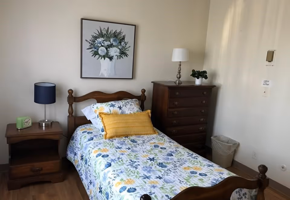 A small bedroom with a single wooden bed covered in a floral bedspread with blue, yellow, and green colors. There is a yellow decorative pillow on the bed. Next to the bed is a wooden nightstand with a blue lamp and a green clock. On the wall above the bed is a framed picture of a white vase with blue and white flowers. To the right of the bed is a tall wooden dresser with a white lamp and a small potted plant on top. A trash can is placed near the dresser against a plain light-colored wall.