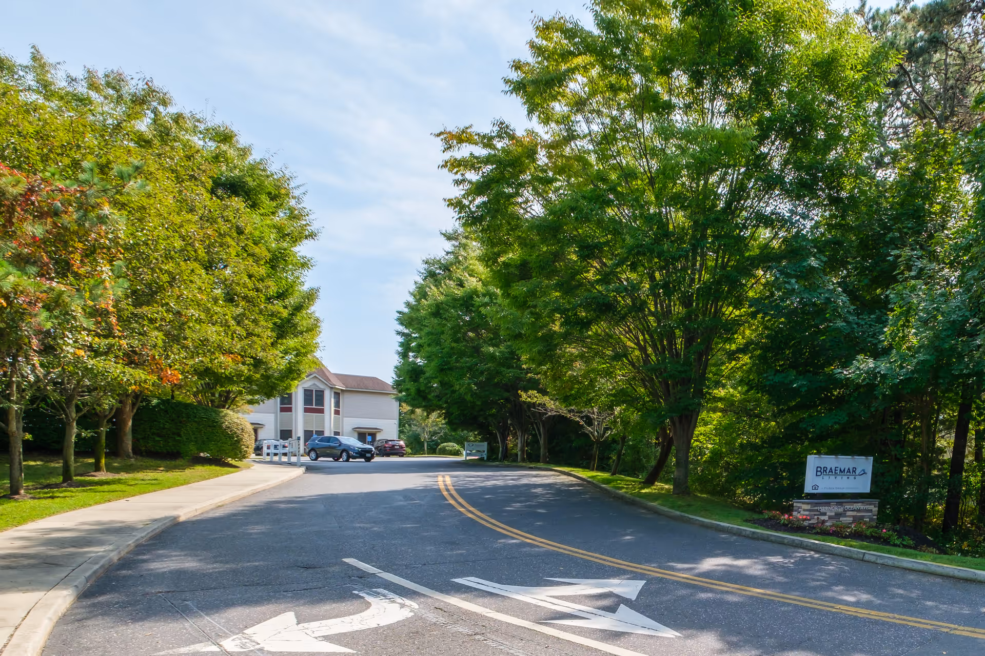 A paved driveway lined with green trees on both sides leading to a two-story building in the background. There is a sidewalk on the left side and a sign on the right side that reads 'Braemar Living'. The sky is clear with some light clouds.