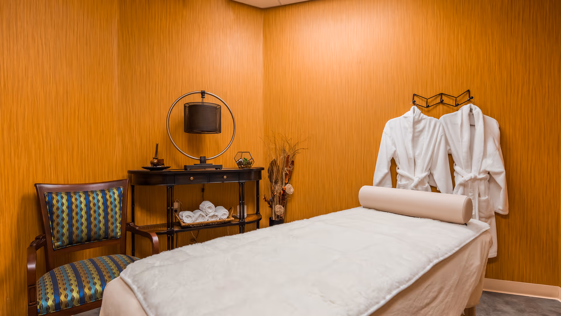 A massage room with a massage table covered in a white sheet and a cylindrical pillow. Two white robes hang on the wall, and there is a wooden chair with a patterned cushion next to a small black table holding rolled white towels and decorative items. The walls are covered in warm wood paneling.