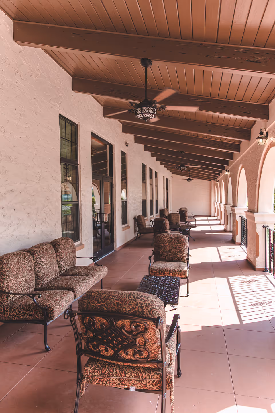 A covered outdoor patio area with cushioned chairs and small tables arranged along the wall. The ceiling has wooden beams and ceiling fans, and the patio is lined with arched openings allowing sunlight to cast shadows on the tiled floor.
