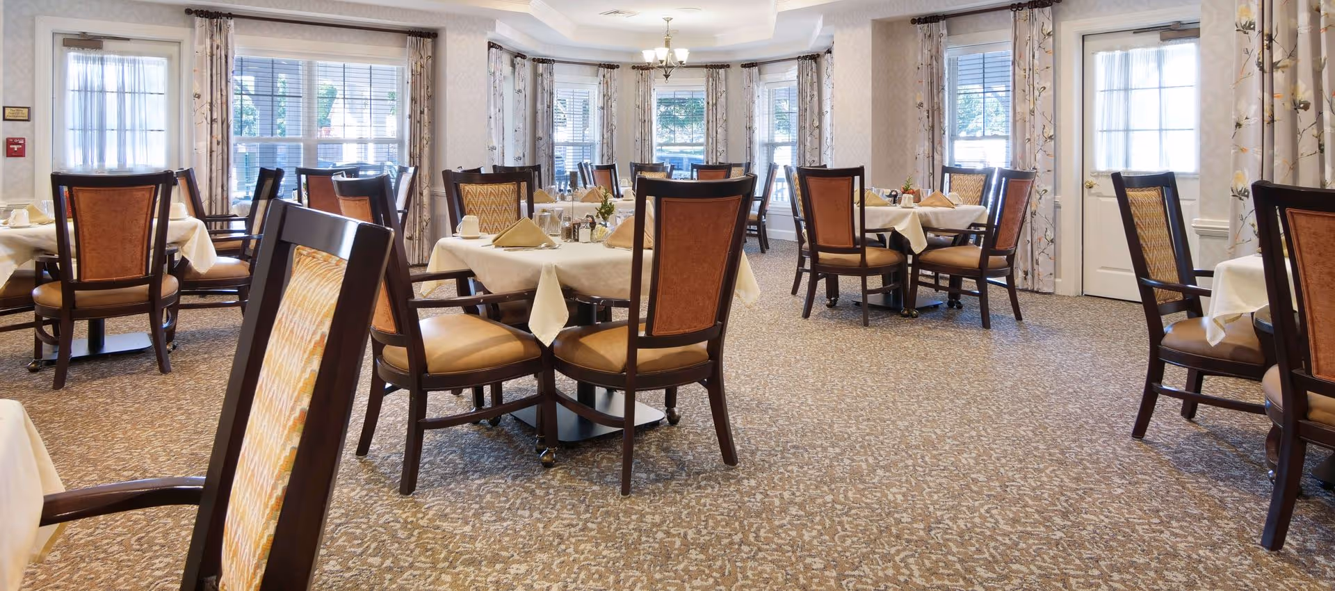 Bright dining room with round tables set with white tablecloths and wooden chairs, large windows and patterned carpet.
