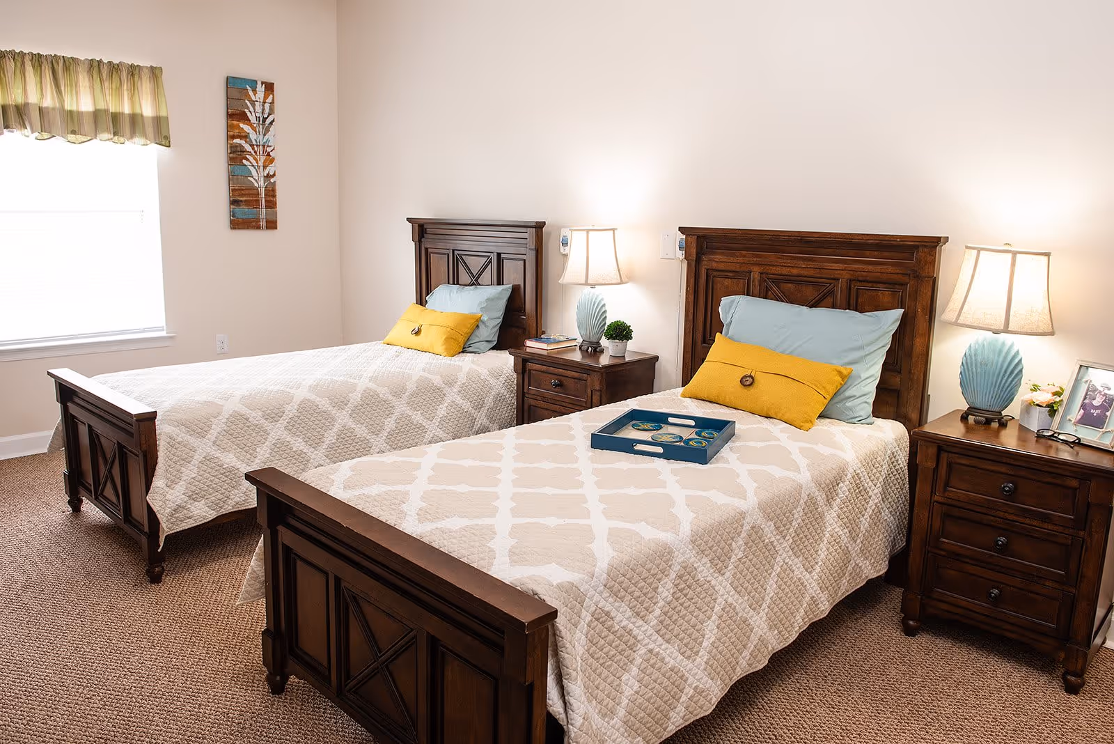 A bright bedroom with two twin beds featuring wooden headboards and footboards. Each bed is made with a beige patterned quilt, a blue pillow, and a yellow accent pillow. Between the beds is a wooden nightstand with a lamp, a small plant, and a few books. Another wooden nightstand with a lamp, a framed photo, and glasses is beside the bed on the right. A window with a green and beige valance lets in natural light, and a decorative wall hanging is on the wall between the window and the beds.