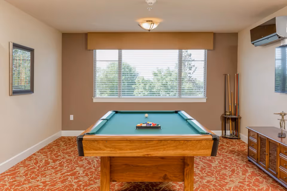 A room with a pool table centered under a window with blinds. The room has beige walls, a patterned carpet, a wall-mounted air conditioner, a wooden cabinet with a decorative item on top, and a rack holding pool cues in the corner.