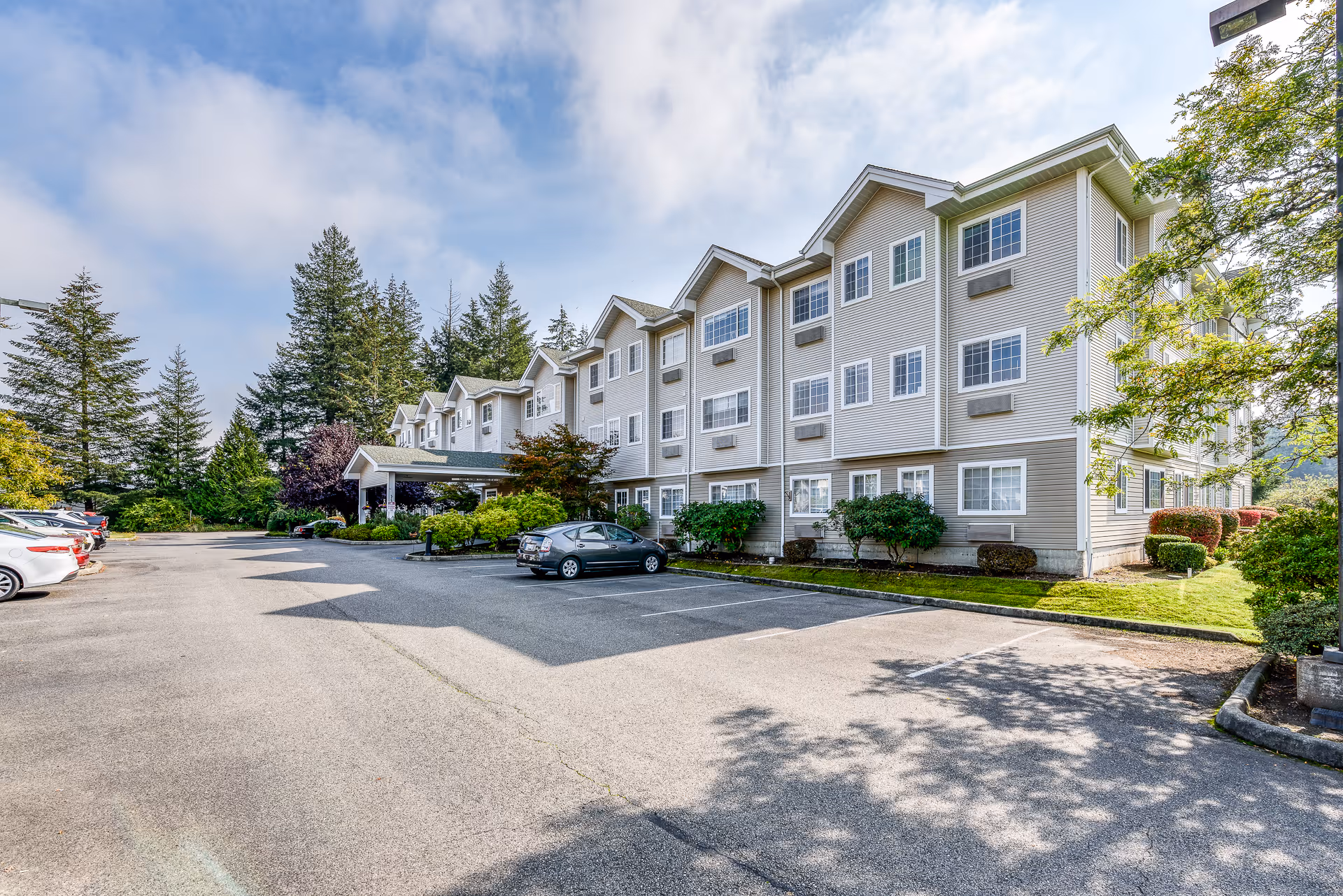 Exterior view of a three-story assisted living facility building with multiple windows, surrounded by trees and greenery. There is a parking lot with several parked cars in front of the building under a partly cloudy sky.