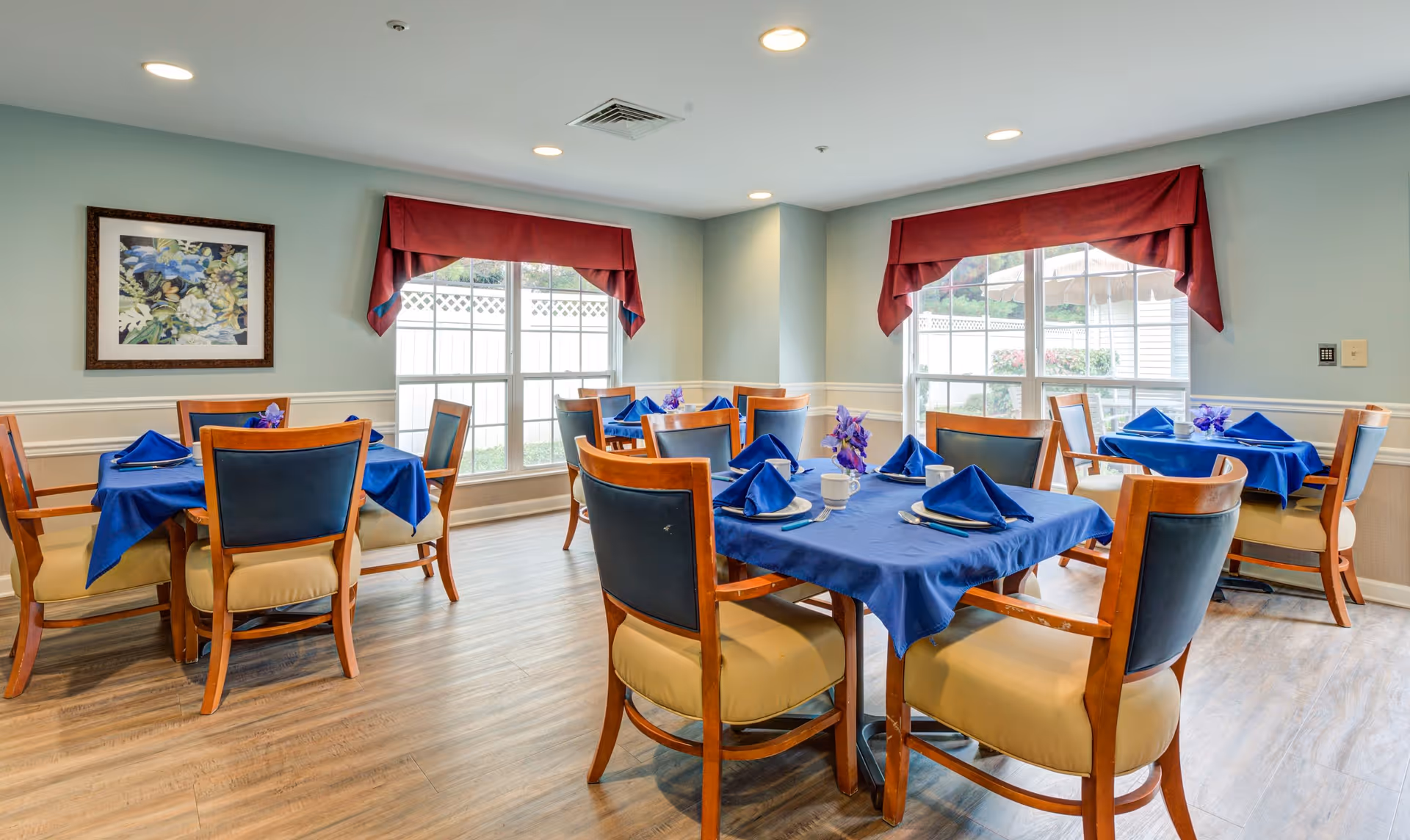 A dining room with several wooden tables covered with blue tablecloths and matching blue napkins neatly folded on each place setting. Each table has wooden chairs with cushioned seats and backs. The room has large windows with red valances, light-colored walls, and wood flooring. A framed floral painting hangs on one wall.