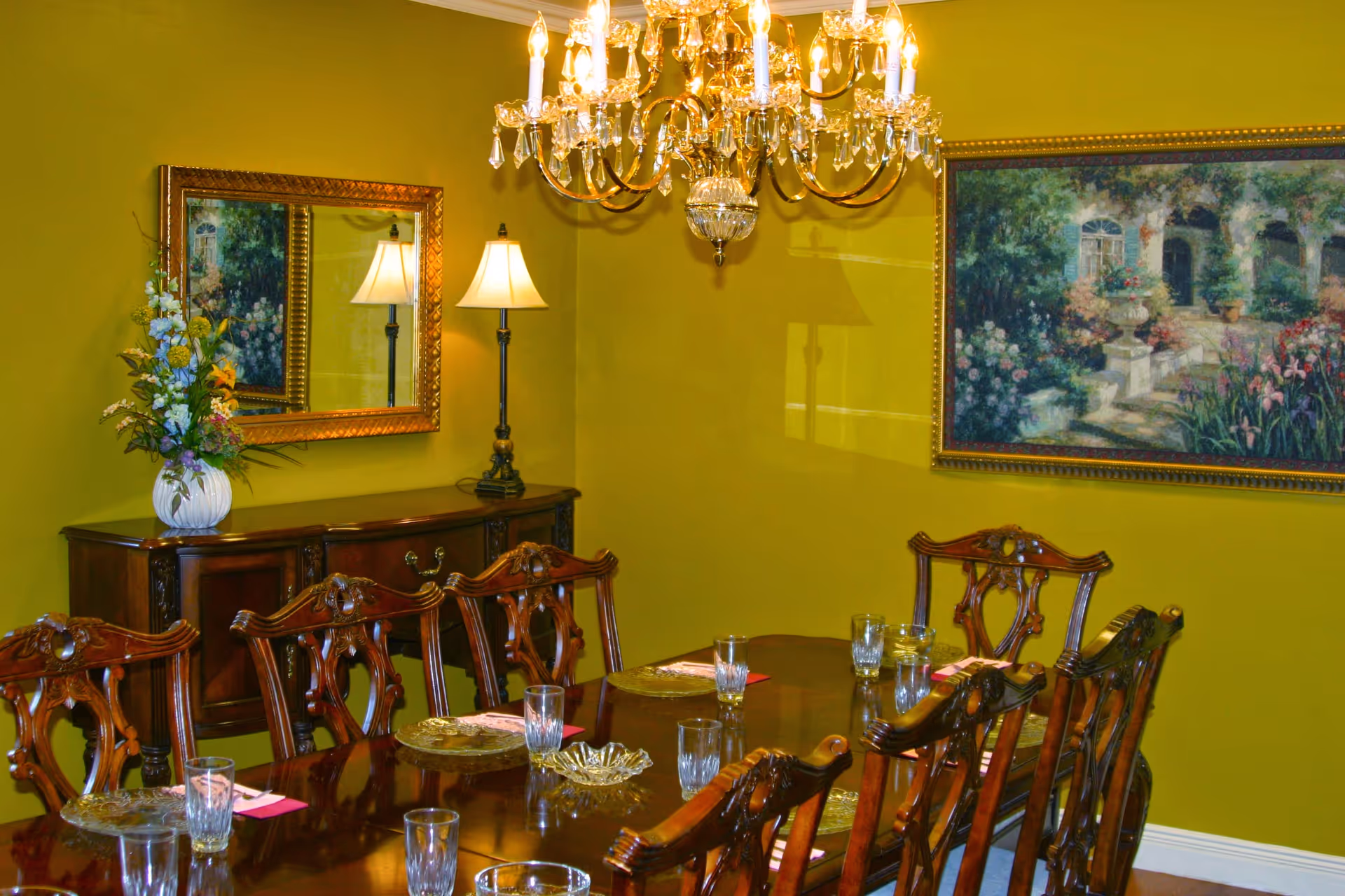 A formal dining room with a long wooden dining table set with glassware and placemats. The room features ornate wooden chairs, a crystal chandelier hanging from the ceiling, a sideboard with a lamp and a vase of flowers, a large mirror, and a framed painting on the wall. The walls are painted green.