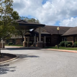Exterior view of a single-story brick building with a covered entrance and a driveway in front, surrounded by trees and shrubs under a partly cloudy sky.