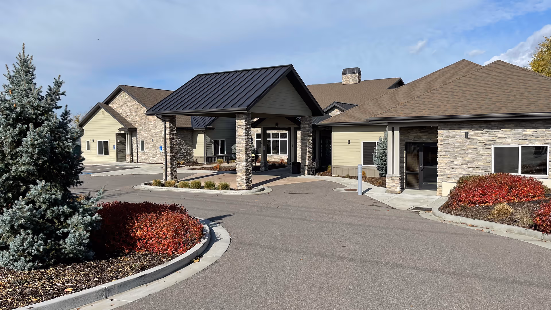 Front entrance and porte-cochère of a single-story assisted living building with stone accents, driveway, and landscaped beds.