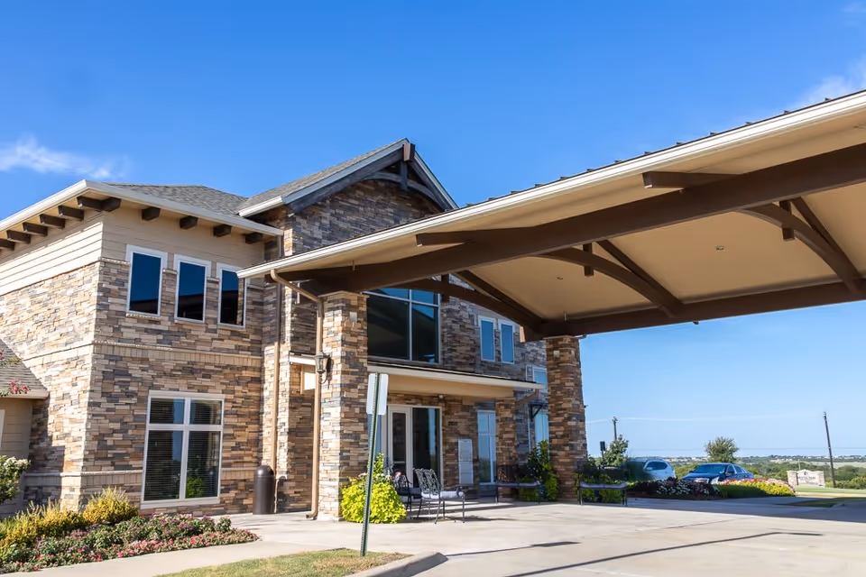Exterior view of a two-story assisted living facility with stone and beige siding, large windows, and a covered entrance driveway. There are some plants and flowers near the building and a clear blue sky above.