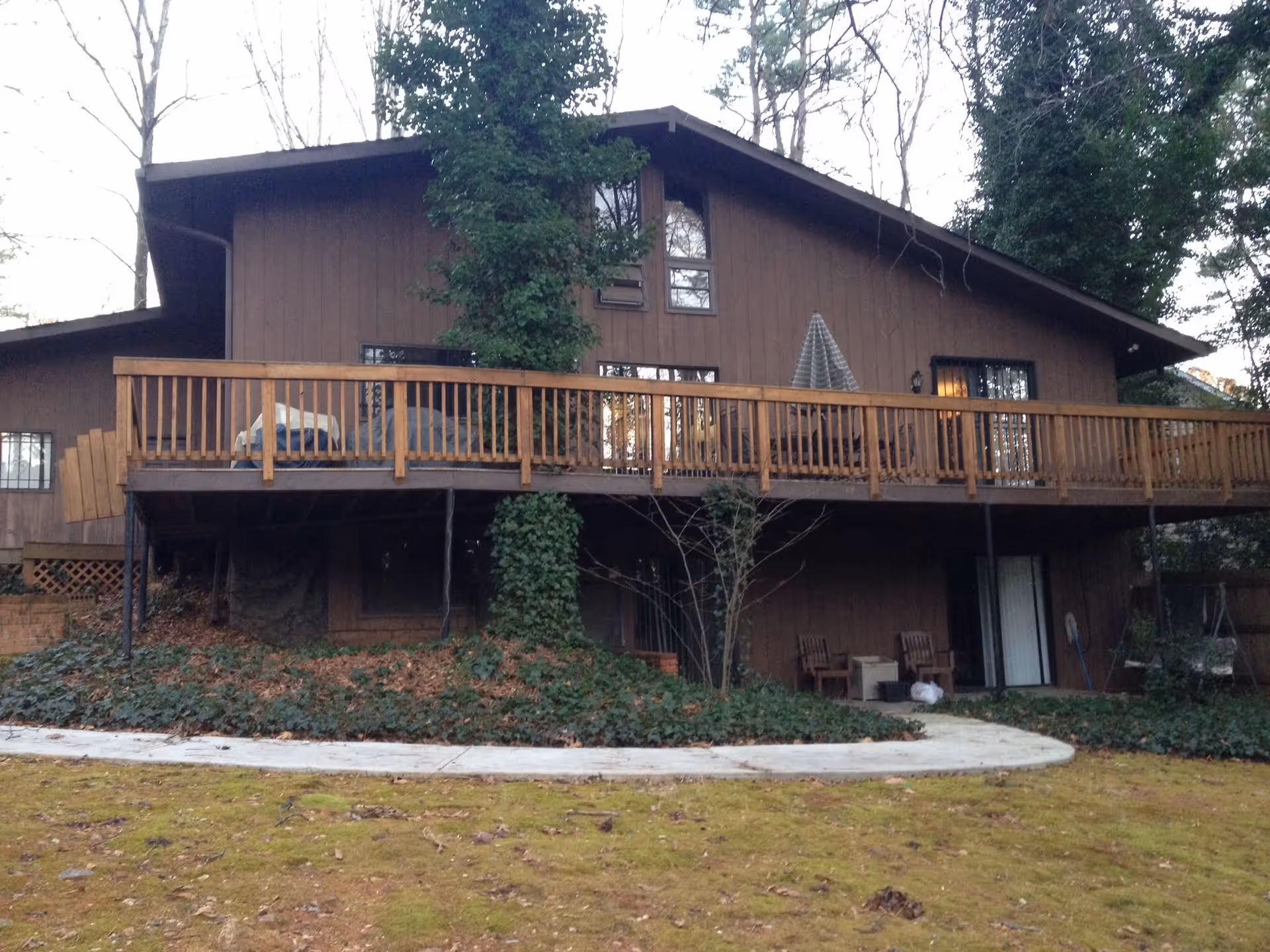 Exterior view of a two-story brown wooden house with a large wooden deck on the upper level. The deck has a railing and outdoor furniture, including a covered grill and an umbrella. The house is surrounded by trees and greenery, with a concrete pathway and a grassy area in the foreground.