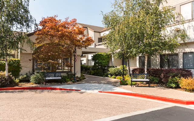 Exterior view of Moraga Royale senior living facility showing the entrance area with a covered walkway, surrounded by trees and shrubs. There are benches on either side of the walkway and a red curb along the driveway.