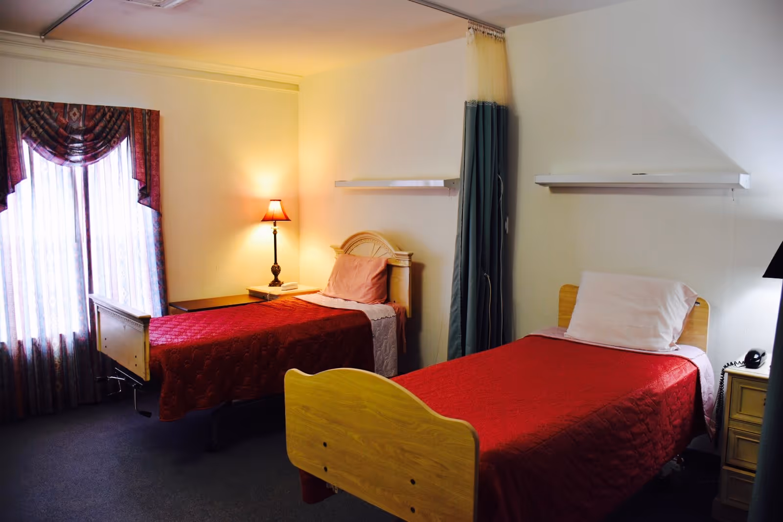 A nursing facility bedroom with two single beds, each covered with red bedspreads and white pillows. There is a nightstand with a lamp between the beds, a window with patterned curtains on the left, and a privacy curtain hanging between the beds.