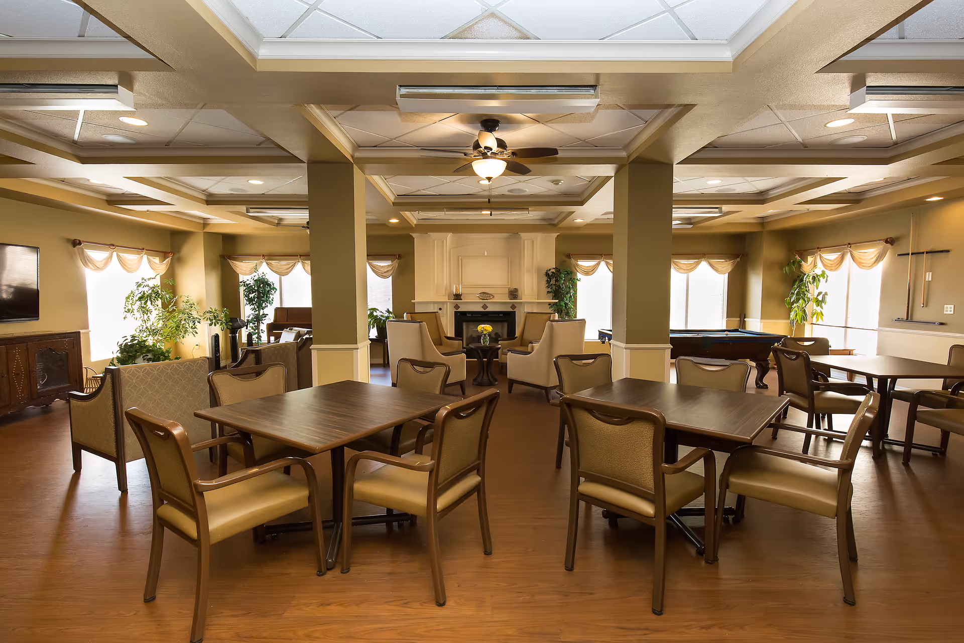 A spacious and well-lit common area in an assisted living facility featuring multiple tables with chairs, comfortable armchairs arranged around a fireplace, large windows with curtains, several green plants, a ceiling fan, and a pool table in the background.