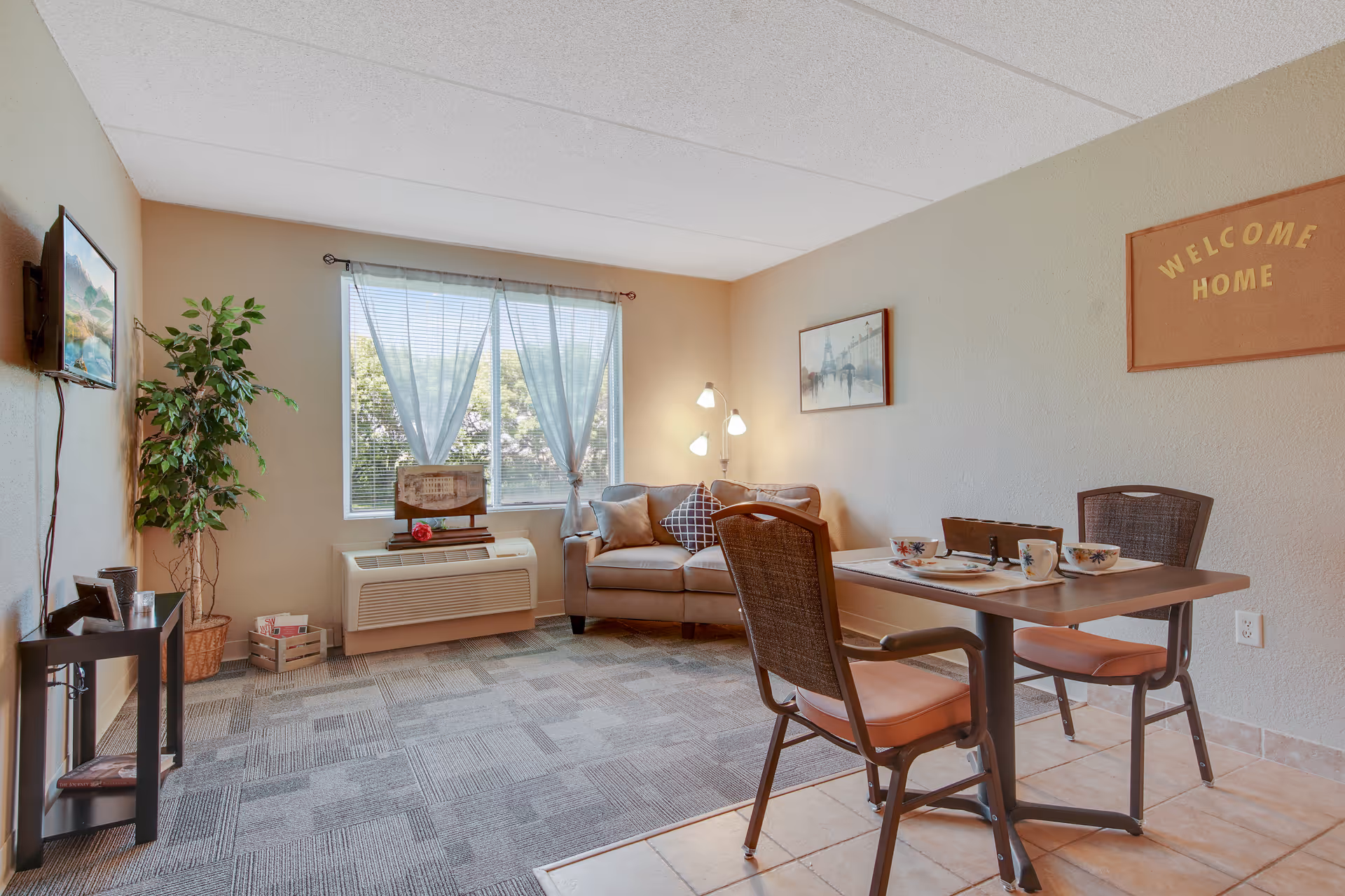 A cozy living room area with a small sofa, a floor lamp, and a window with sheer curtains letting in natural light. There is a small dining table with two chairs set with cups and plates. A potted plant is placed near the wall-mounted TV, and a framed picture and a 'WELCOME HOME' sign hang on the walls. The floor is a combination of carpet and tile.