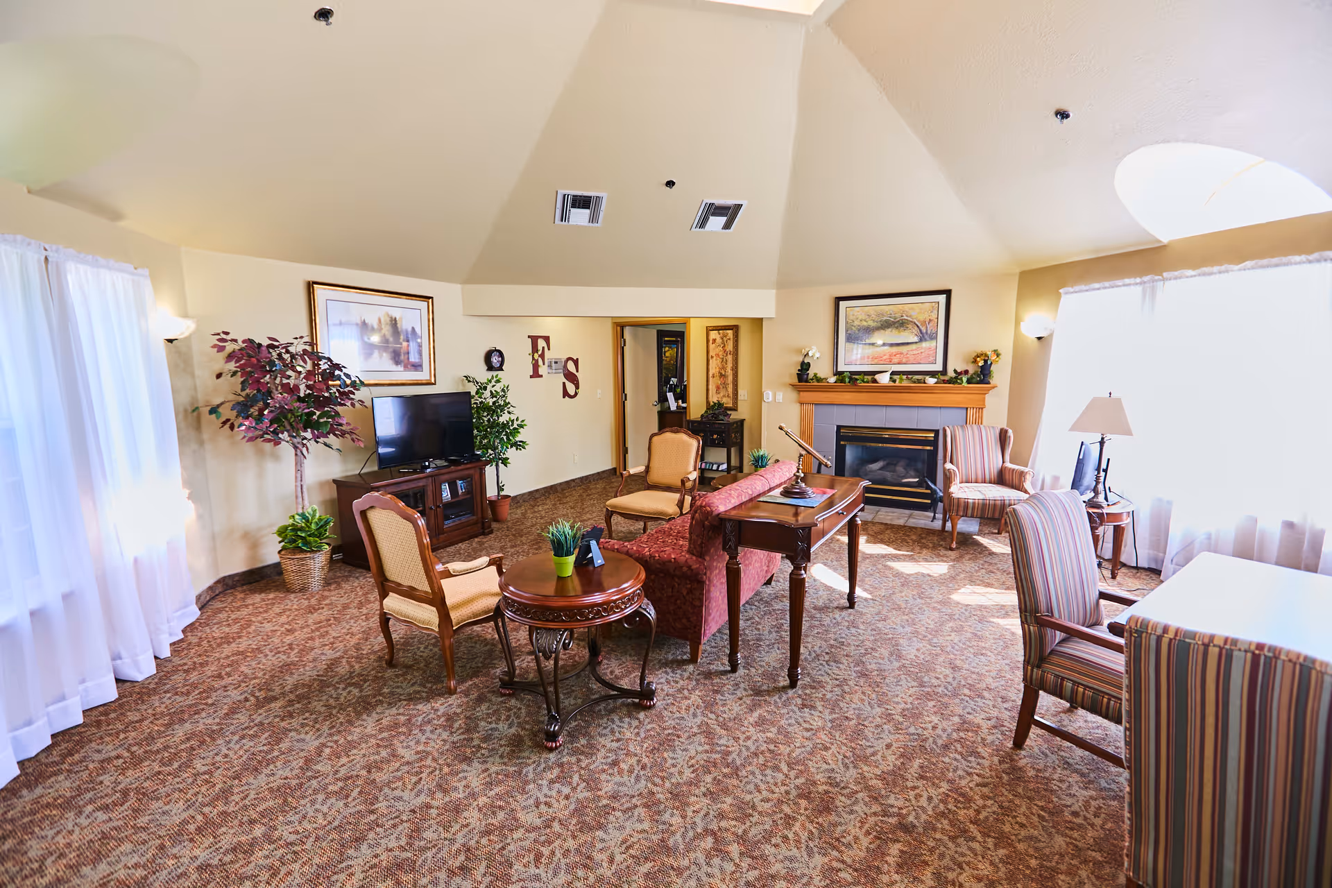 A cozy living room with a patterned carpet, a red upholstered sofa, several wooden chairs with cushions, a wooden coffee table, and a fireplace with a painting above it. There is a TV on a stand, potted plants, and large windows with sheer white curtains allowing natural light to fill the room.