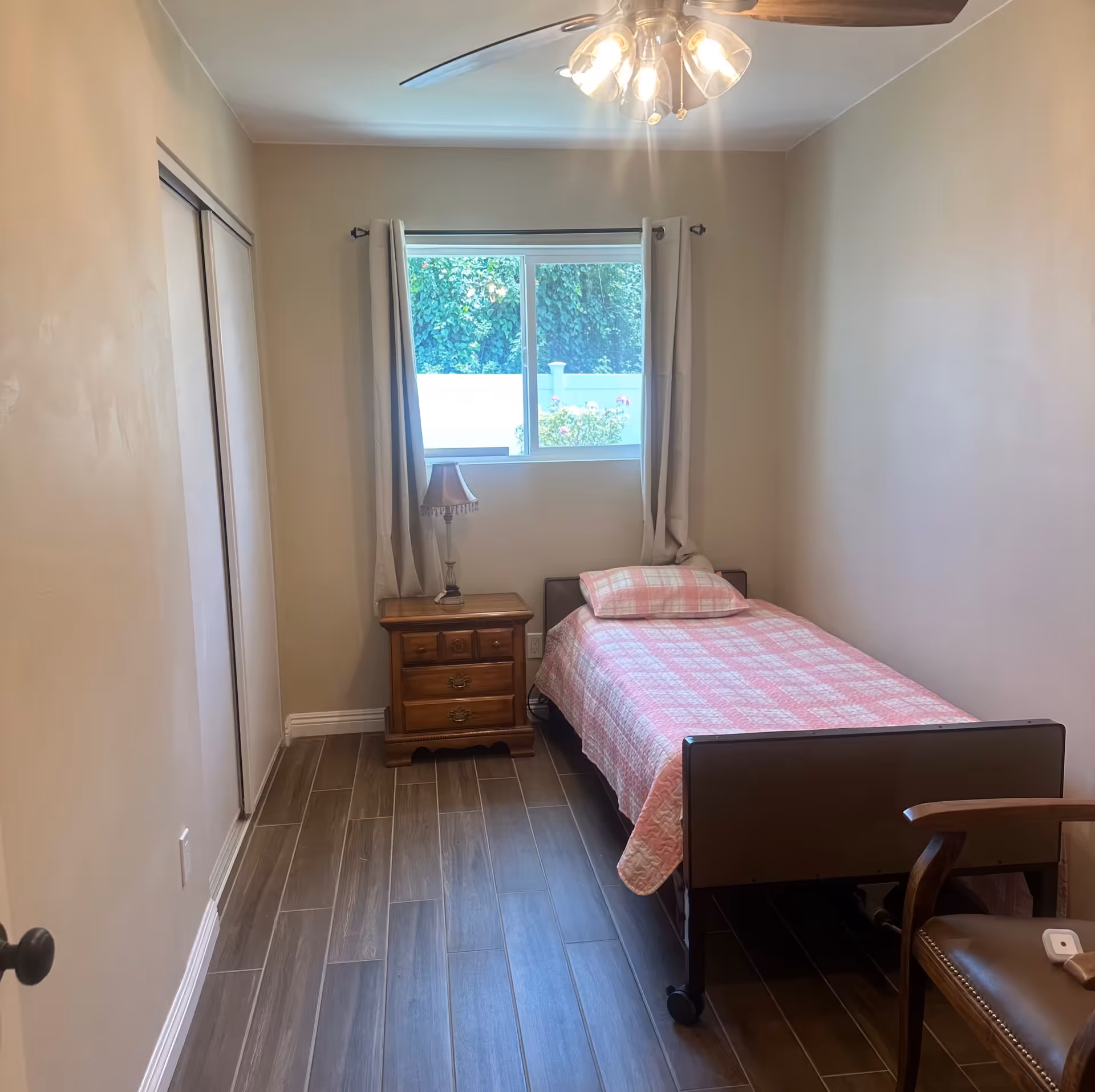 A small, simple bedroom with a single bed covered in a pink checkered quilt and matching pillow. Next to the bed is a wooden nightstand with a lamp. A window with beige curtains lets in natural light, and there is a wooden chair with a cushion in the corner. The floor is covered with wood-like tiles and the walls are painted beige.