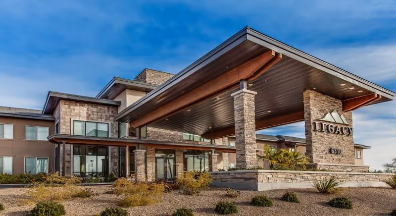Exterior view of Legacy House of Centennial Hills, a modern senior living facility with stone and stucco facade, large covered entrance supported by stone pillars, and landscaped desert plants in front under a blue sky.