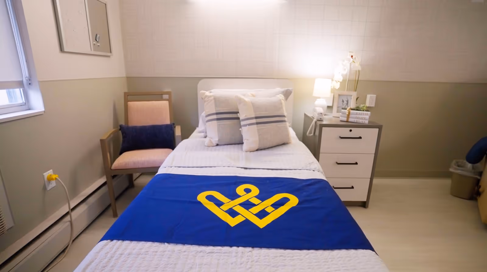 A neatly made single bed with white bedding and two pillows, covered partially by a blue blanket with a yellow heart-shaped design. Next to the bed is a small nightstand with a lamp, books, a framed photo, and a small plant. A beige armchair with a dark blue cushion is positioned beside the bed near a window with a white blind. The room has light-colored walls and flooring, creating a calm and clean atmosphere.