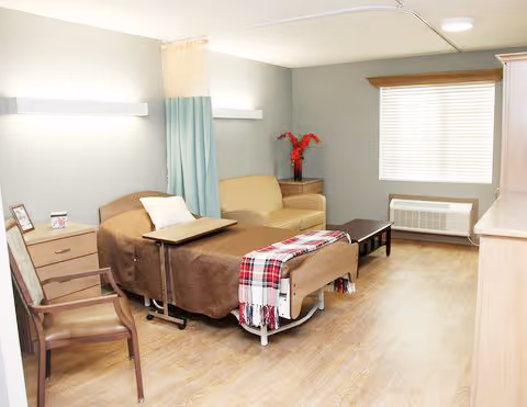 A bright senior-care room with an adjustable bed, bedside table and tray, armchairs, a small sofa, coffee table, and a window with blinds.