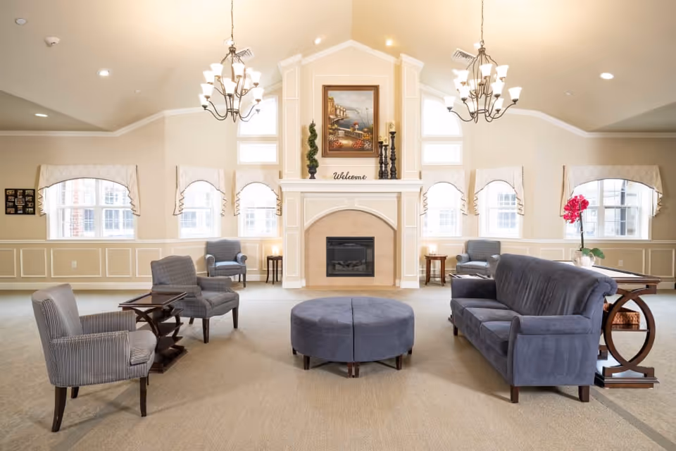 Bright communal lounge with a central fireplace, chandeliers, and arranged sofas, chairs, and a round ottoman.