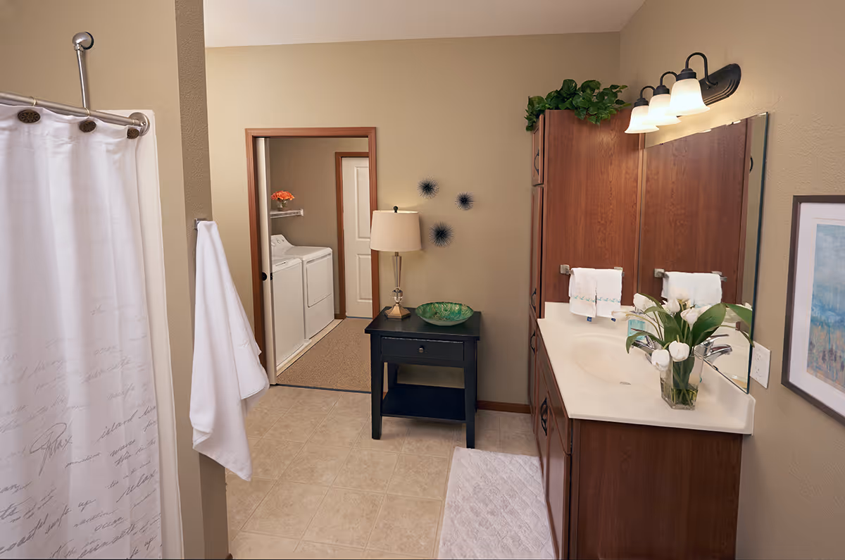 A bathroom with a shower curtain on the left, a white towel hanging on the wall, a black side table with a lamp and decorative bowl, and a wooden vanity with a sink, mirror, and flowers. In the background, there is a laundry area with a washer and dryer.