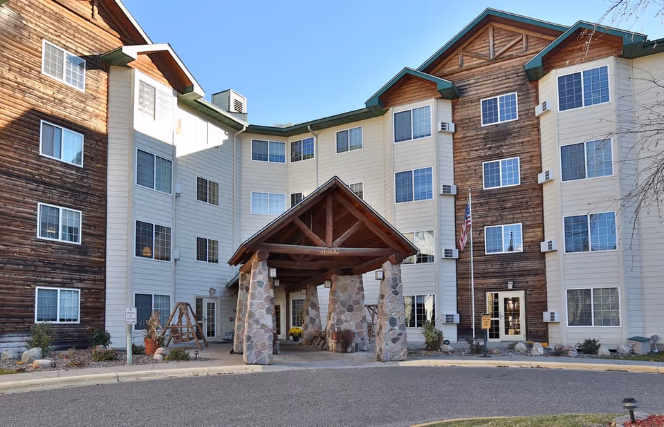 Front exterior view of a multi-story senior living facility with a covered entrance supported by stone pillars. The building features a combination of wood and light-colored siding with multiple windows and an American flag near the entrance.