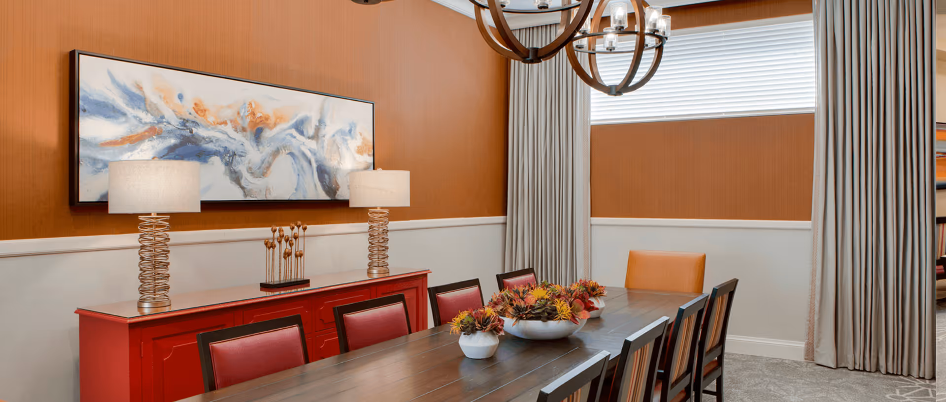 A dining room with a long wooden table surrounded by chairs. On the table are decorative bowls with artificial plants. Against the wall is a red sideboard with two lamps and a modern abstract painting above it. The walls are painted orange and white, with a window covered by beige curtains and a chandelier hanging from the ceiling.