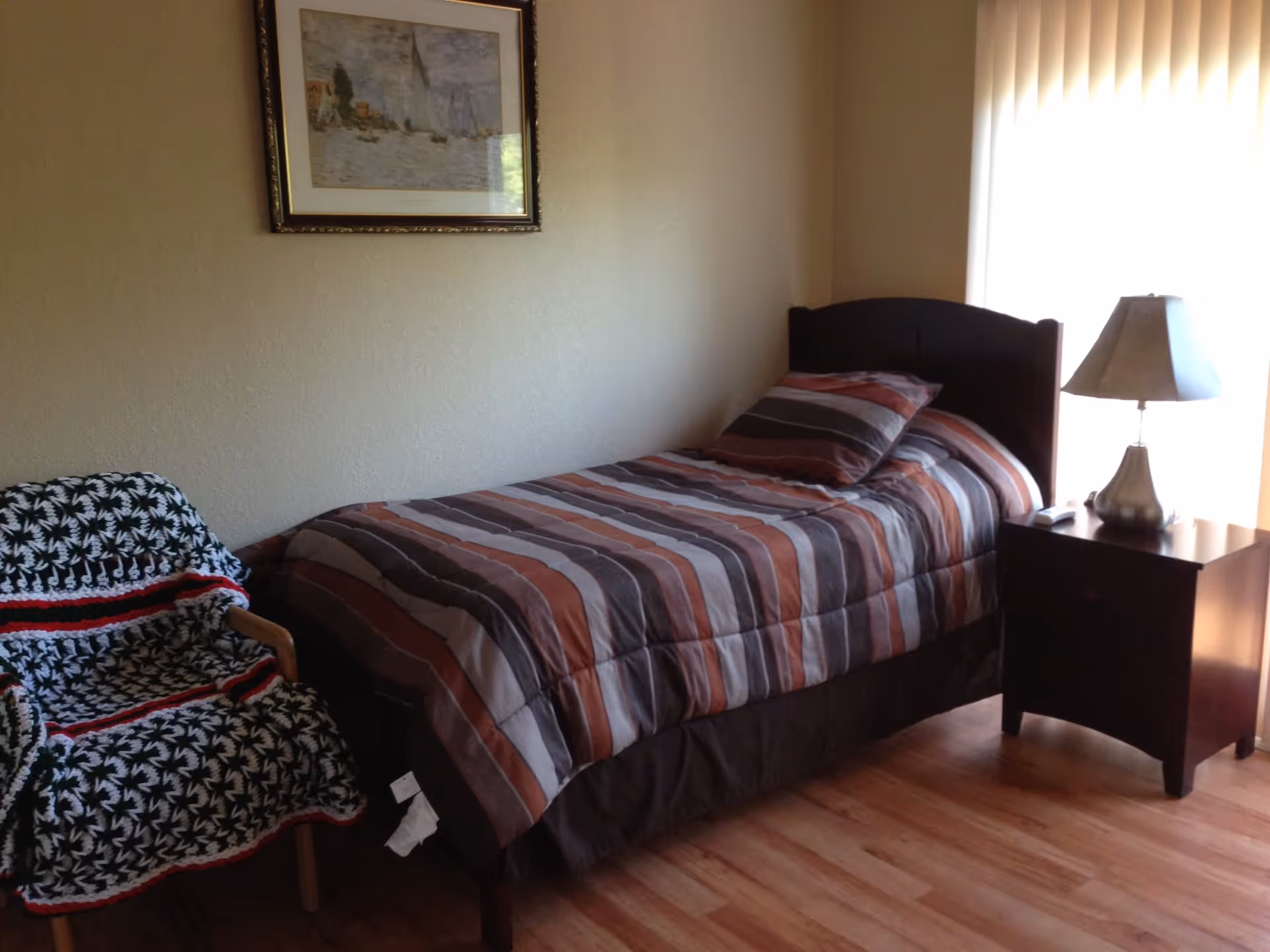 A small bedroom with a single bed dressed in striped bedding, a nightstand with a lamp, and a chair covered by a blanket.