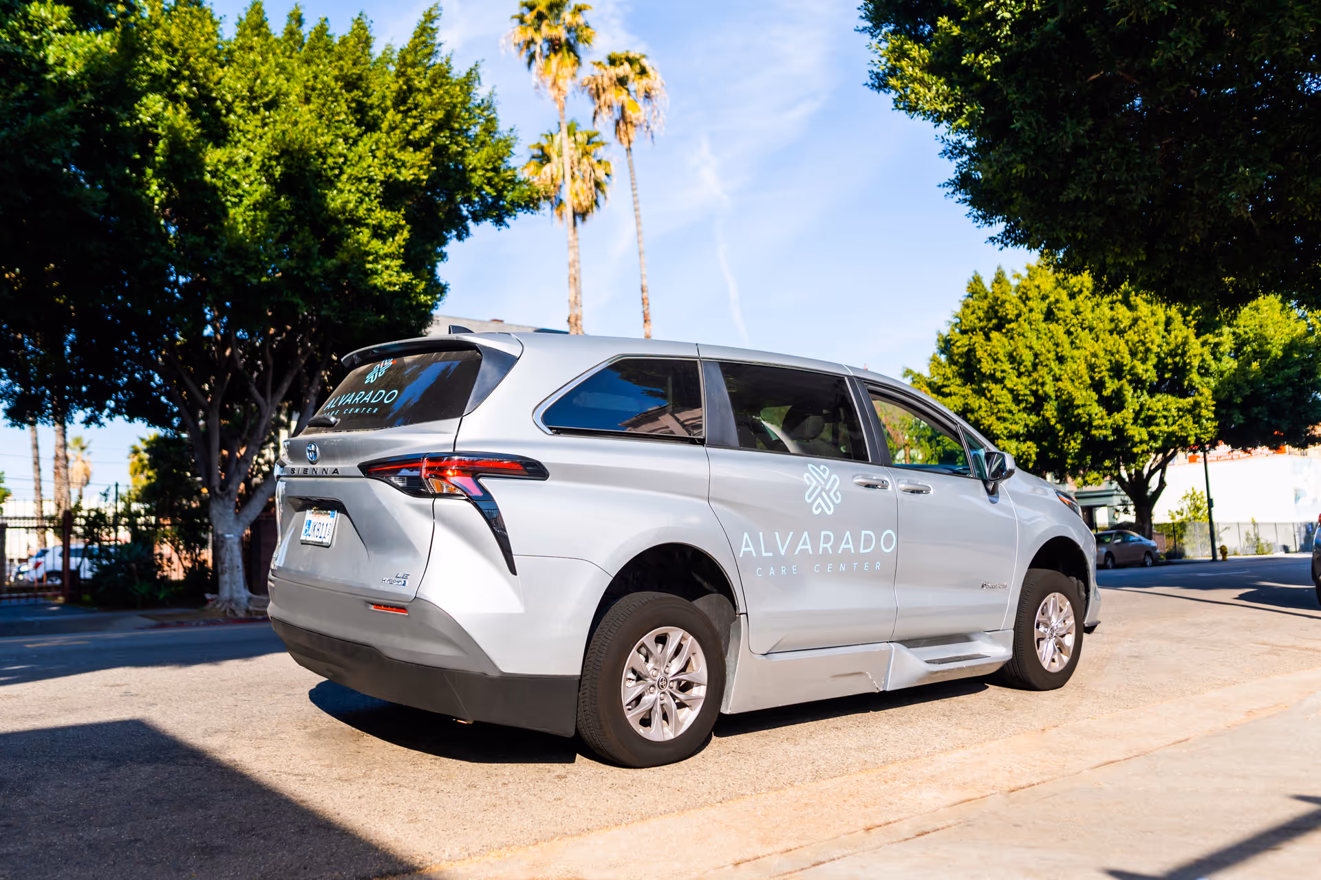 Silver minivan parked on a street with the logo and text 'Alvarado Care Center' on the side. The background includes trees, a fence, and a clear blue sky with palm trees.