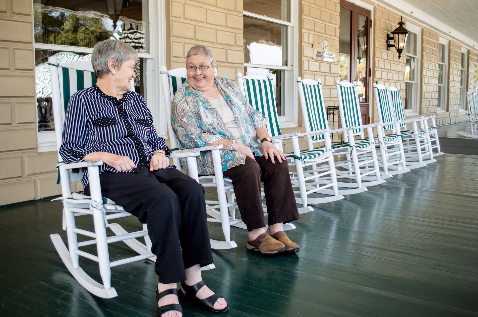 Two elderly women sitting and chatting on white rocking chairs with green and white striped cushions on a covered porch with a green floor and beige siding walls.