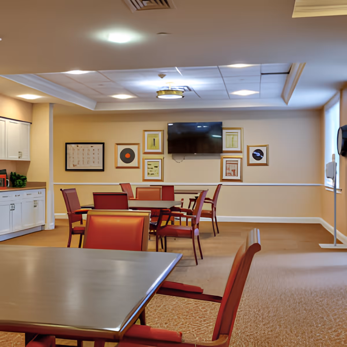 A well-lit common room with several tables and red cushioned chairs arranged around them. The walls are beige with framed artwork and a flat-screen TV mounted on one wall. There is a kitchenette area with white cabinets on the left side and large windows on the right side letting in natural light.