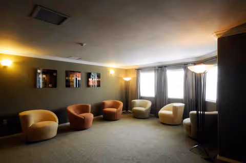 A cozy lounge area with six modern, rounded armchairs arranged in a semi-circle along the walls. The room has soft lighting from wall sconces and floor lamps, three abstract art pieces on the olive green wall, and three large windows with curtains letting in natural light.