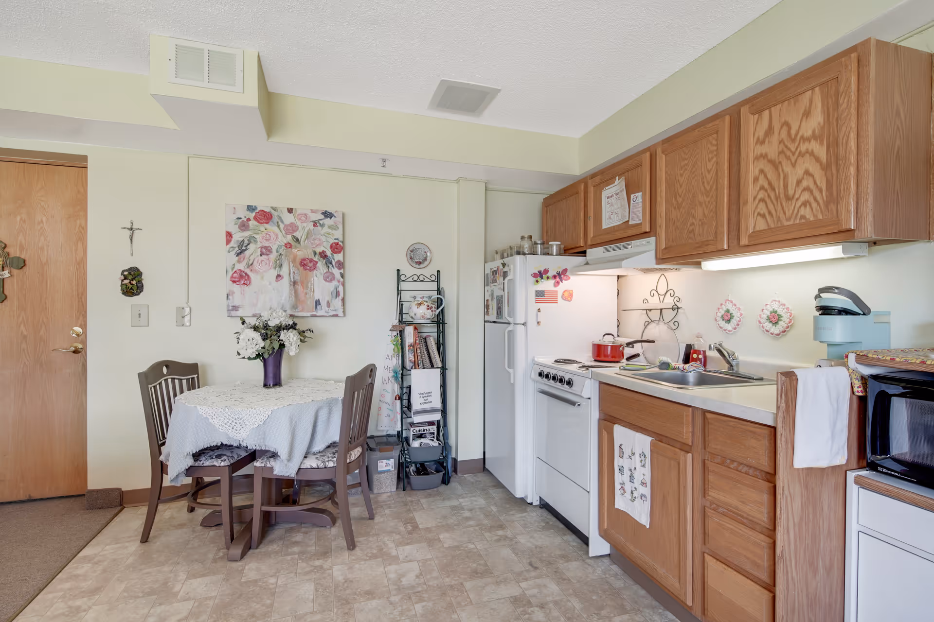 A small kitchen and dining area in an assisted living facility. The kitchen has wooden cabinets, a white refrigerator, a stove with a red pot on it, a sink, and a microwave. The dining area features a round table covered with a lace tablecloth and two wooden chairs. There is a floral painting on the wall above the table, a small metal rack with books and kitchen items, and a wooden door to the left.