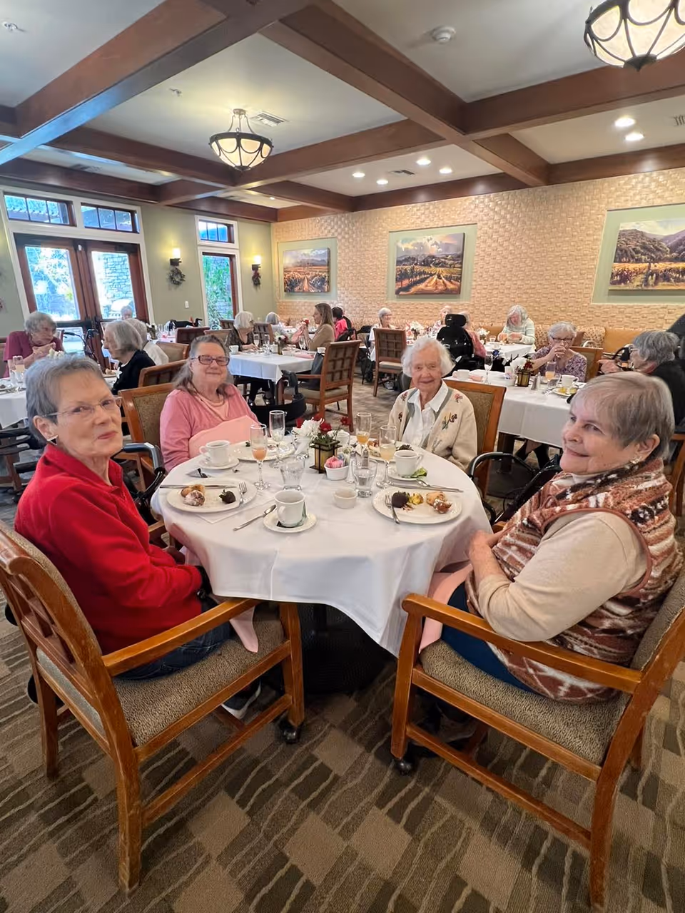 A group of elderly women sitting around a round dining table in a well-lit dining room at Atria Valley View. The table is set with plates, cups, glasses, and small desserts. Other elderly residents are visible dining in the background. The room has wooden beams on the ceiling, framed landscape paintings on the walls, and large windows letting in natural light.