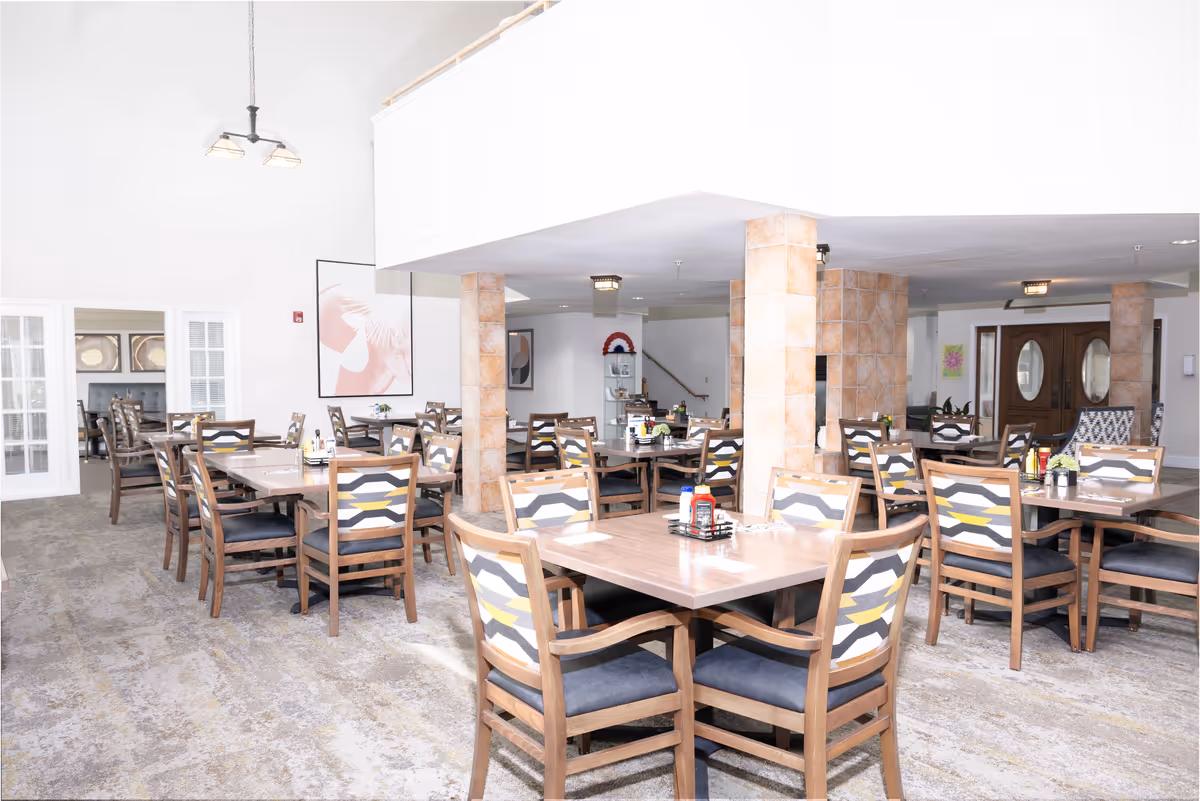 A bright and spacious dining room in a senior living facility with multiple wooden tables and chairs arranged neatly. The chairs have patterned upholstery with shades of gray, white, and yellow. The room features tiled columns, light-colored walls, and a carpeted floor. There are framed artworks on the walls and a chandelier hanging from the ceiling.
