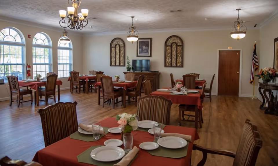 A dining room with multiple tables covered in red tablecloths, each set with plates, glasses, napkins, and small floral centerpieces. The room has wooden floors, large arched windows letting in natural light, chandeliers hanging from the ceiling, and an American flag near a closed wooden door. There is a TV on a wooden cabinet against the far wall, flanked by decorative wall hangings.