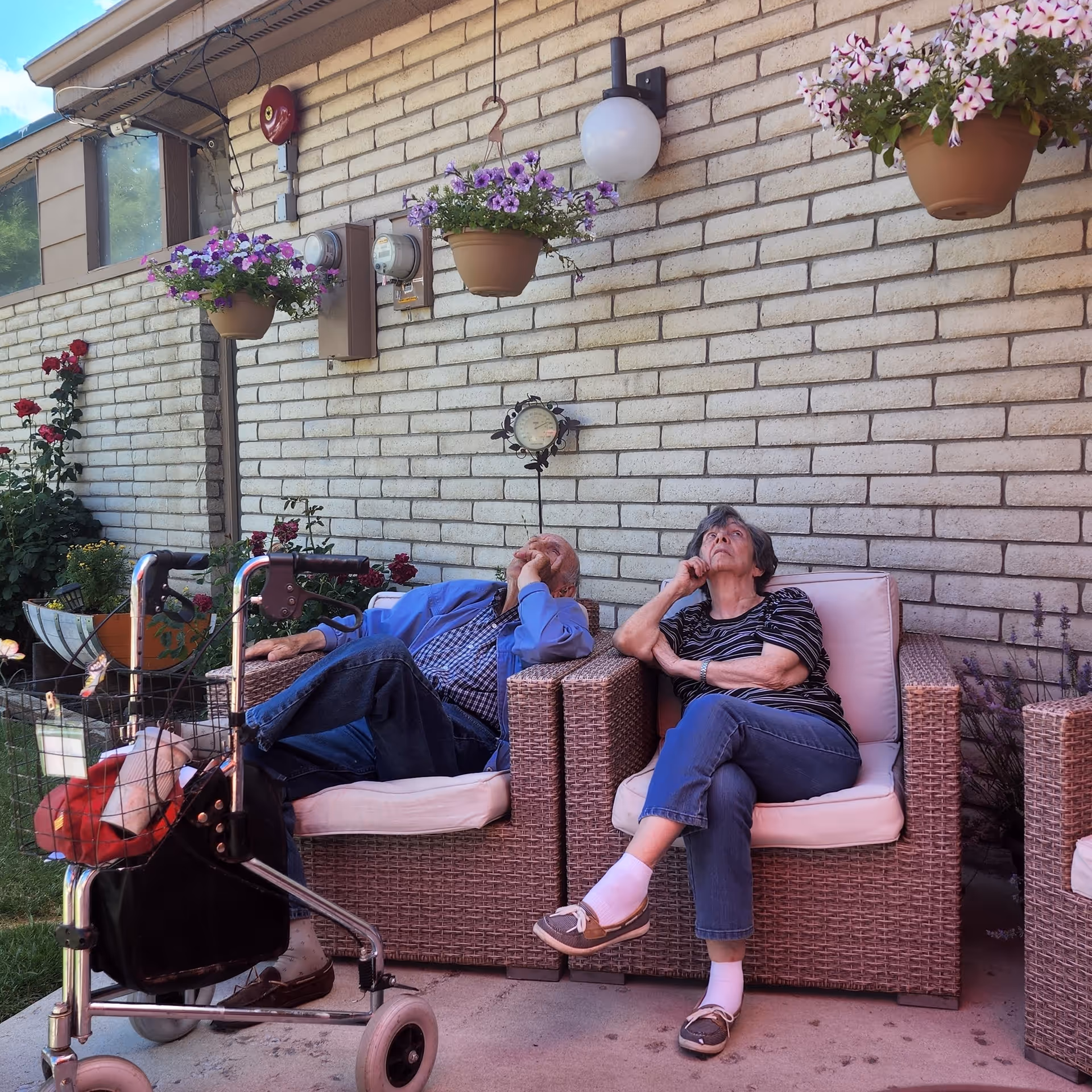 Two elderly individuals sitting on cushioned wicker chairs outside against a brick wall. There are hanging flower pots with purple flowers above them and a walker with a basket in front of one person. The setting appears to be a garden or patio area with some greenery and flowers nearby.