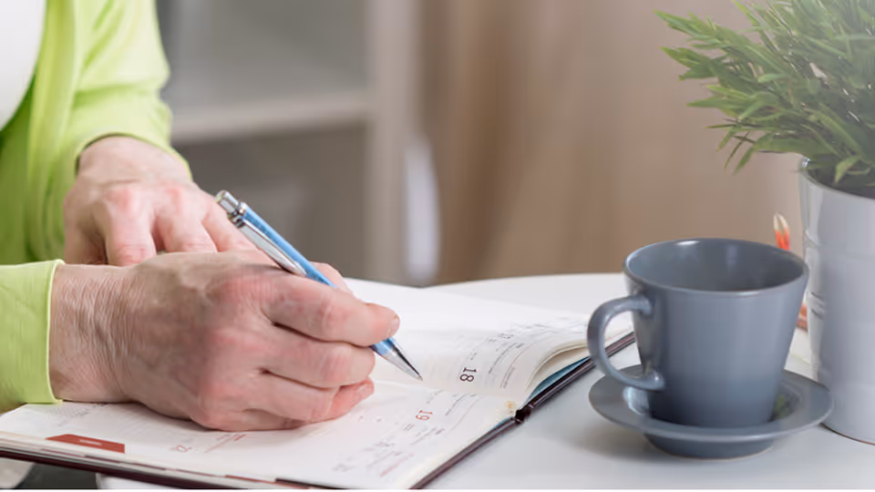 Close-up of an elderly person's hands writing in a planner or calendar with a blue pen on a white table. Next to the planner is a gray cup and saucer, and a small potted plant in a white pot.
