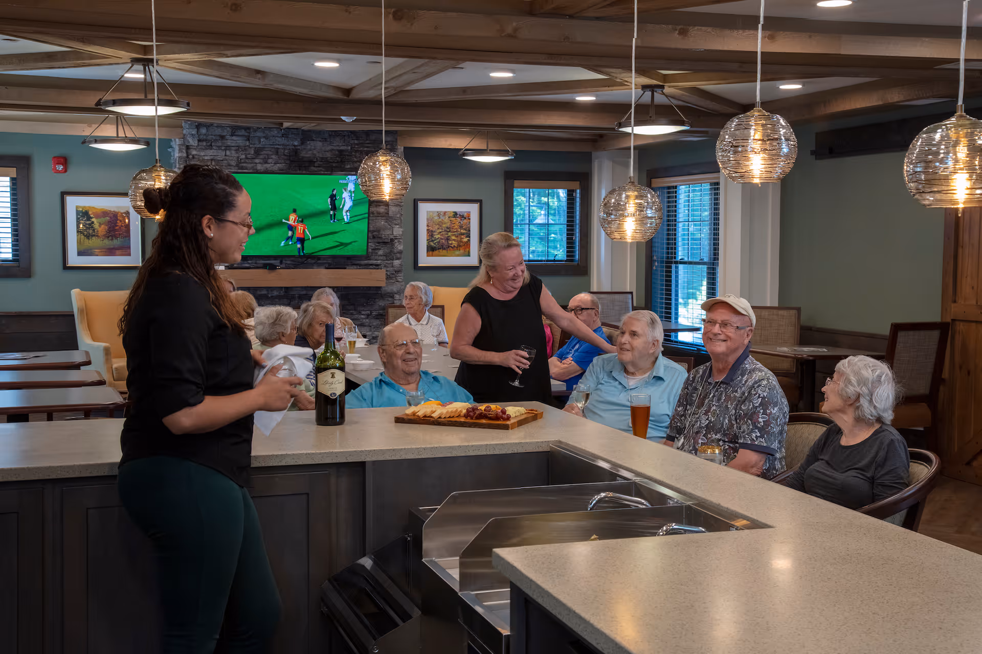 A group of elderly people sitting and socializing around a counter in a well-lit common area with pendant lights. A woman stands behind the counter holding a wine bottle and a cloth, while another woman is standing and engaging with the seated group. A TV mounted on a stone wall shows a soccer game, and framed artwork is visible on the walls.