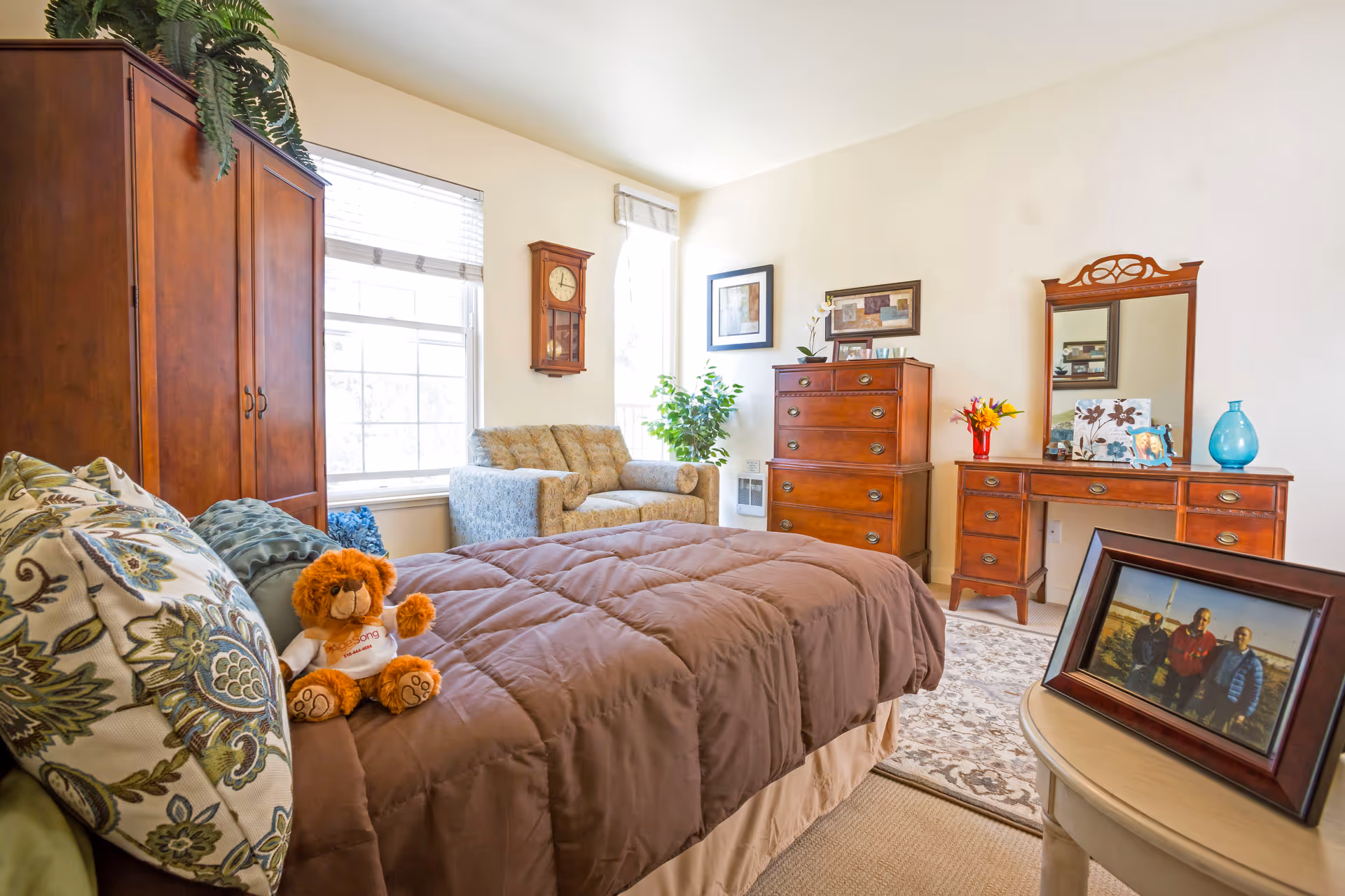A cozy bedroom with a brown comforter on the bed, decorative pillows, and a small teddy bear wearing a white shirt. The room features wooden furniture including a wardrobe, a chest of drawers, and a dresser with a mirror. There is a patterned armchair near two large windows letting in natural light. A round side table holds a framed photo of three people. The walls are decorated with framed artwork and a wooden wall clock.